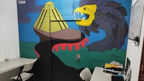 Ein Klassenzimmer mit Wandgemälde, das eine Pyramide und einen Dinosaurier zeigt. Es sind Tische und Stühle vorhanden, sowie eine Tafel an der Wand.