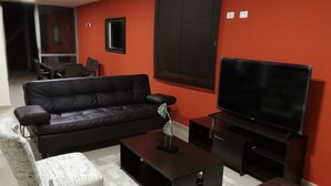 Uma sala de estar com um sofá de couro preto, uma poltrona branca com estampa, uma mesinha e uma televisão de tela plana grande em frente a uma parede vermelha.