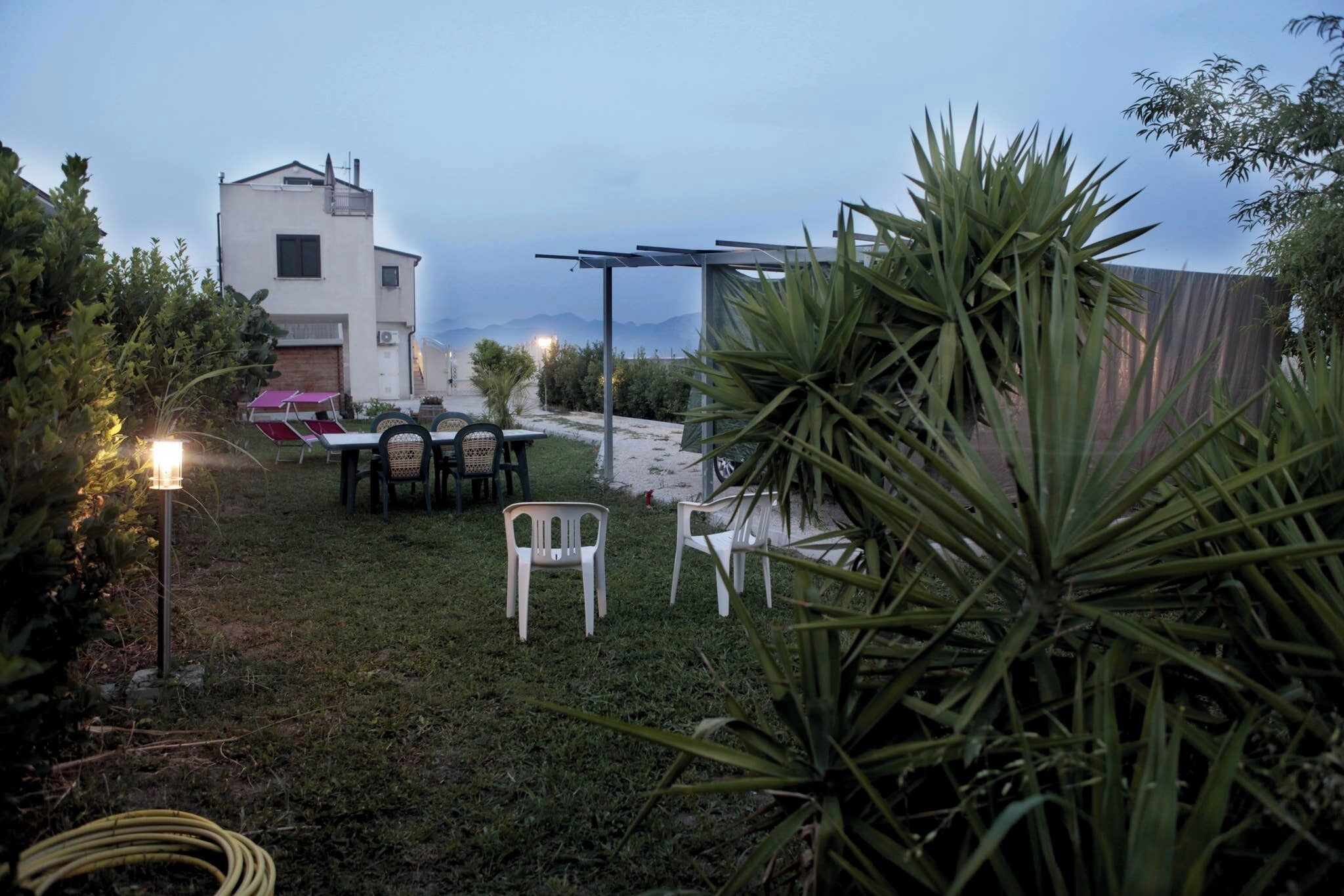 Ein grünes, offenes Areal mit Tisch und Stühlen, einem Gebäude und einer Pergola mit Beleuchtung.