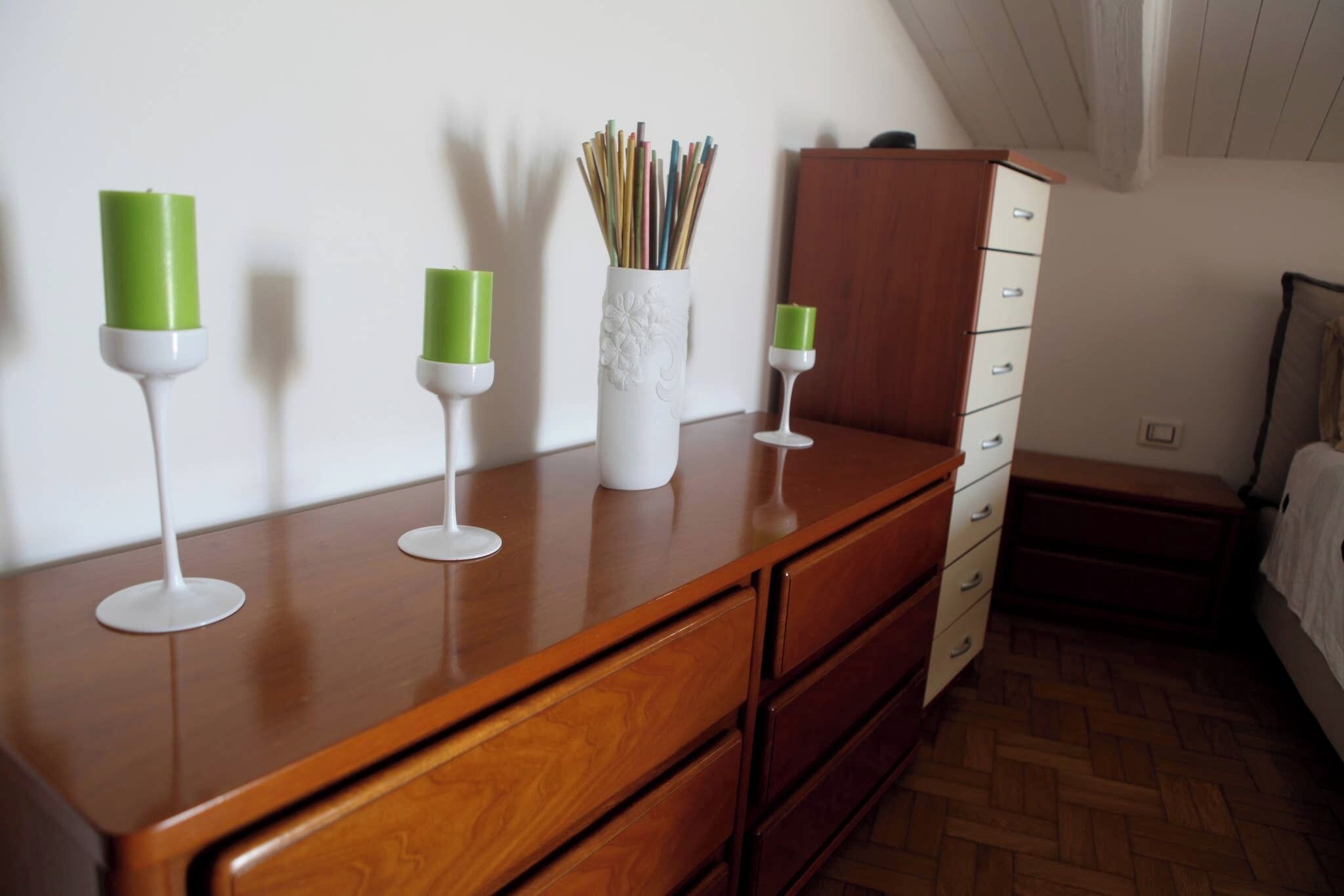 Une commode en bois avec des tiroirs, un vase blanc orné de motifs floraux et trois bougeoirs verts placés sur le dessus.
