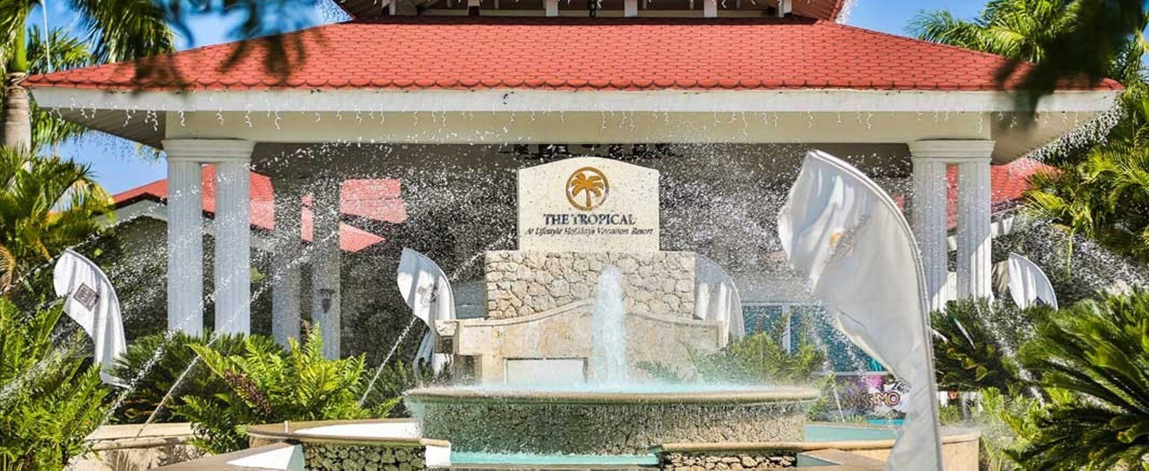 A water fountain with a sign reading 'The Tropical' in front of a building with a red roof.