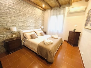 Une chambre avec un mur en pierre, un lit avec des oreillers et une couverture, une table de chevet, une commode et une fenêtre avec des rideaux.