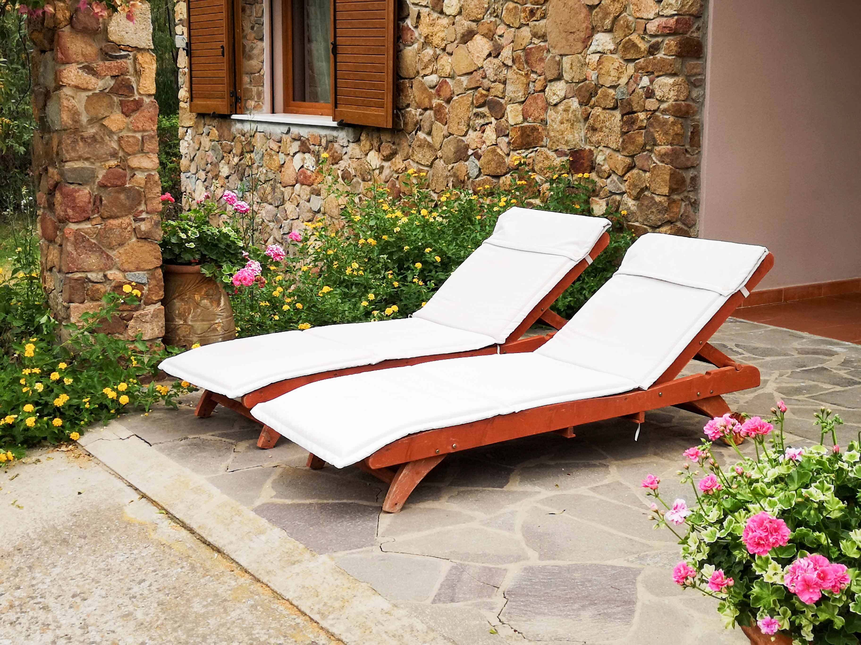 Two wooden lounge chairs with white cushions, positioned on a stone patio with a stone wall and a window with wooden shutters in the background.