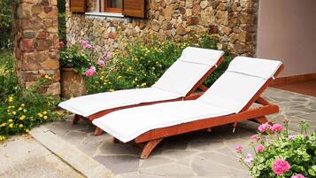 Two wooden lounge chairs with white cushions, positioned on a stone patio with a stone wall and a window with wooden shutters in the background.