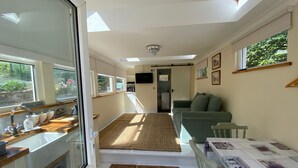 A compact, well-lit room with a kitchenette, a sofa, a dining table, and a large window providing natural light.