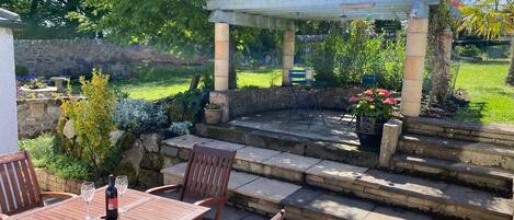 A patio with a wooden table and chairs, a pergola, and a garden with various plants and a stone wall.