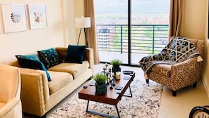Una sala de estar con sofá, sillón y mesa de centro, un ventanal con vistas, y obras de arte enmarcadas en la pared.