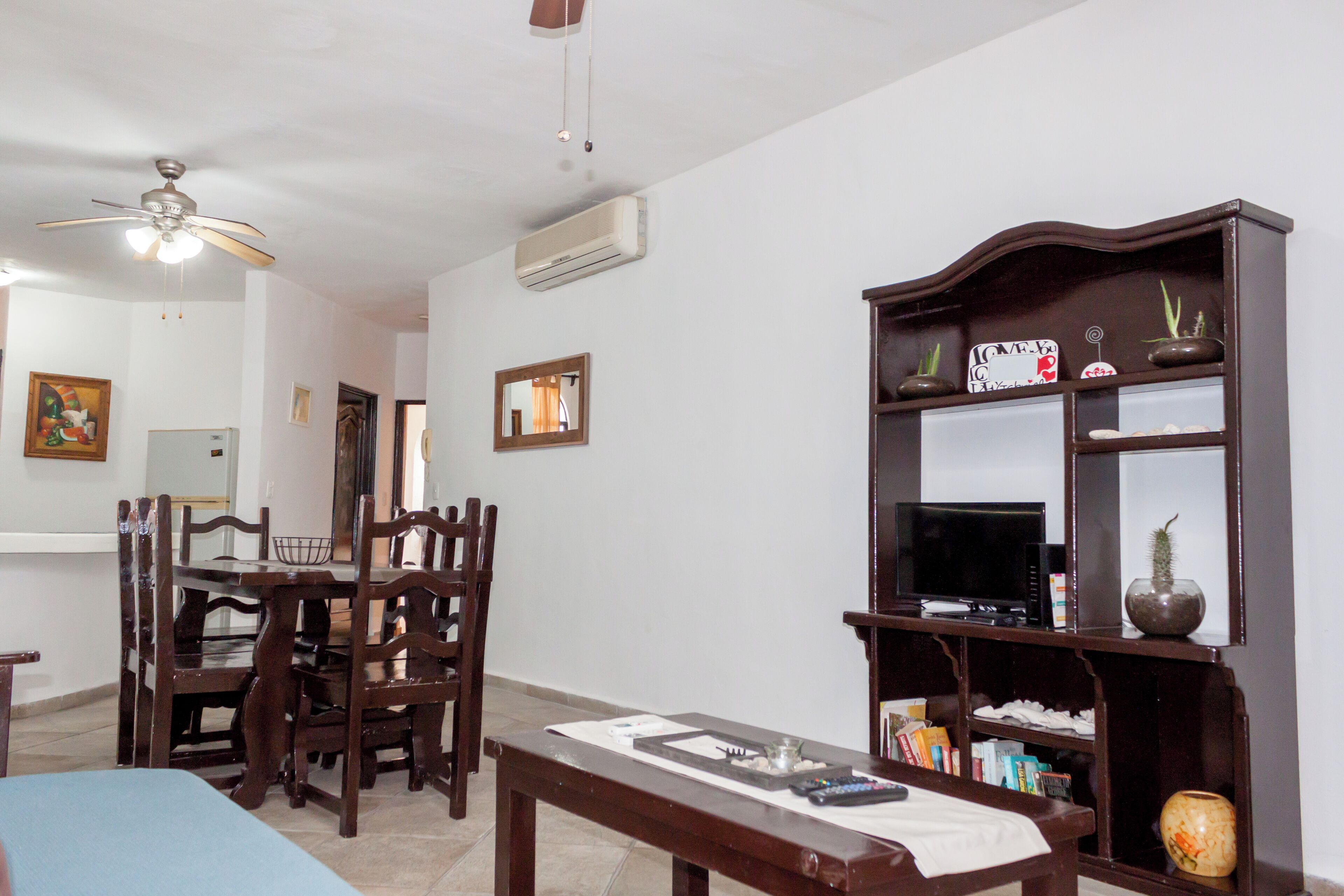 Una sala de estar con comedor, ventilador de techo, aire acondicionado, televisor y estantería con objetos decorativos.