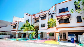 Un hôtel à plusieurs étages, doté d’une façade blanche et de bordures rouges, avec des balcons, une cour intérieure agrémentée de palmiers et un espace de stationnement.