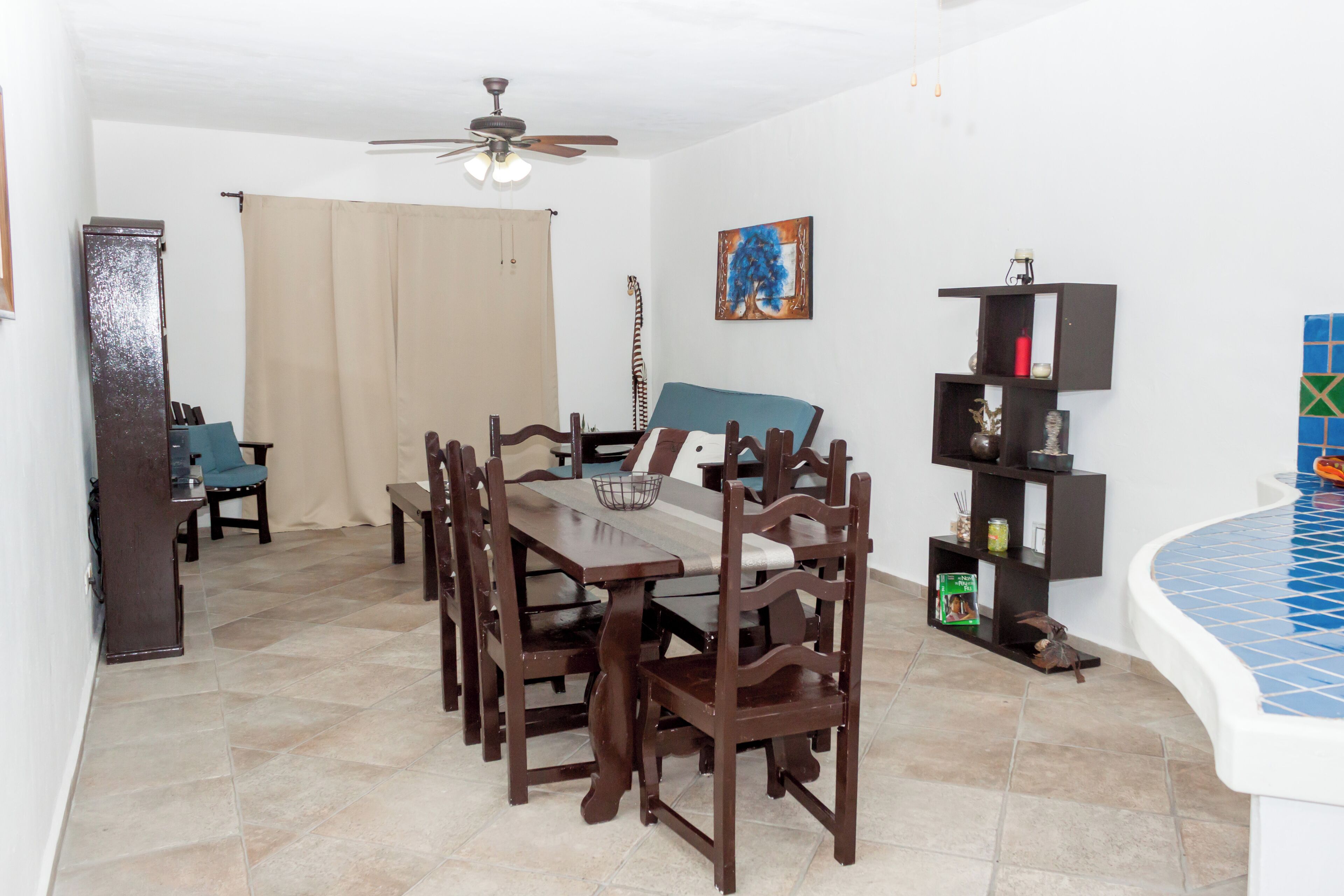 Un coin repas avec une table et des chaises en bois, un ventilateur de plafond, une chaise bleue, une guitare et une étagère avec des objets décoratifs.