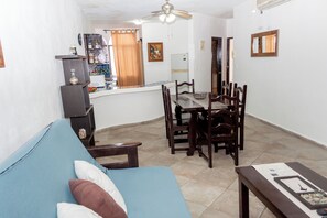 Ein Wohnzimmer mit blauem Sofa, einem Esstisch mit Stühlen, einem Deckenventilator und einer Küche im Hintergrund.