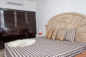 Ein Hotelzimmer mit kariertem Bettbezug, einem Holzkopfende mit floralen Schnitzereien, einem weißen Nachttisch mit Lampe und dunklen Holzschränken.