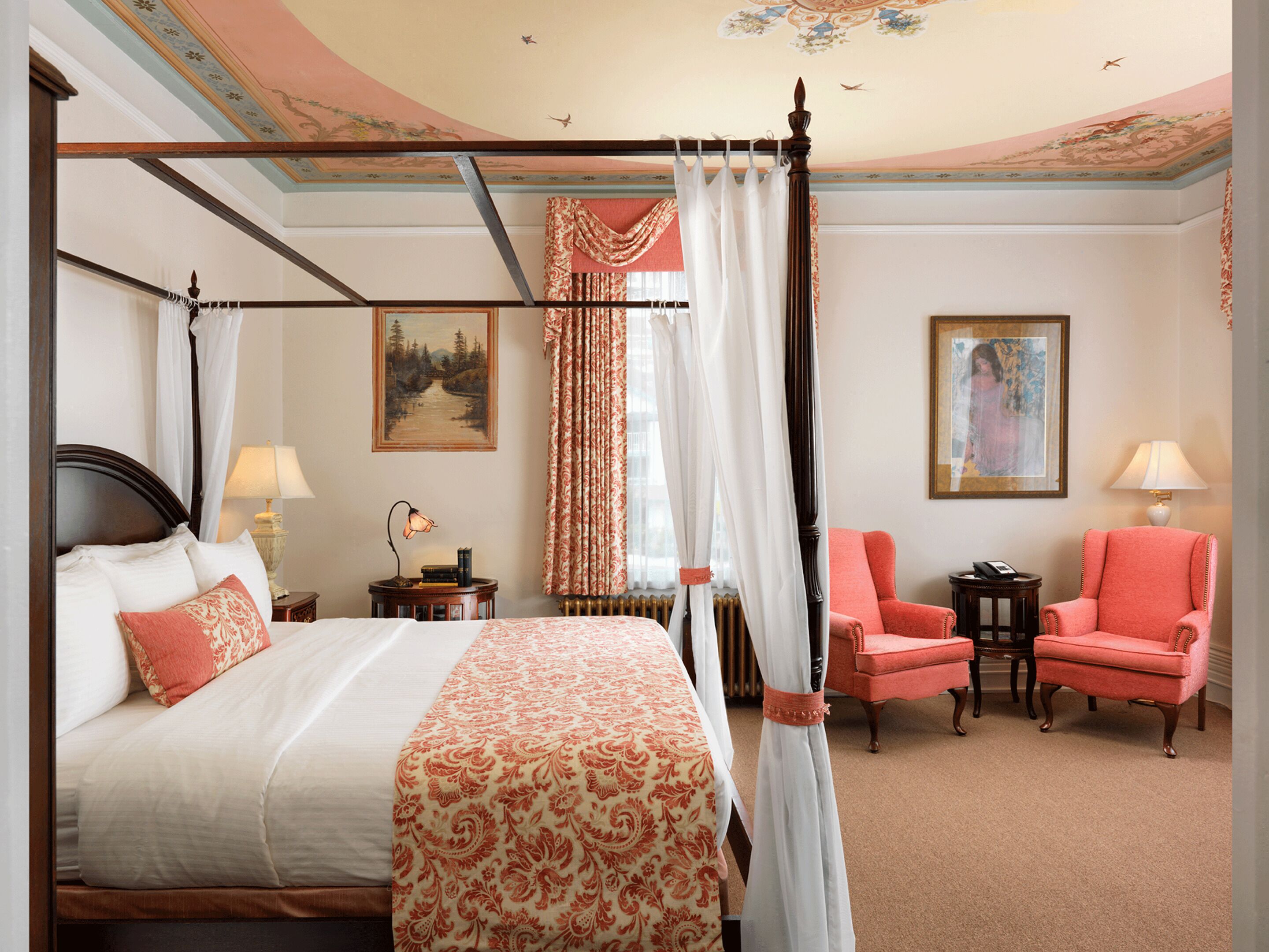 A four-poster bed with a canopy, a sitting area with two chairs, a small table, and a painting on the wall.