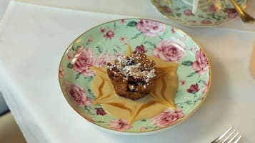 Eine blumengeschmückte Teekanne gießt Tee in eine passende Tasse und Untertasse, ein Dessertteller und eine Gabel auf einem Tisch.