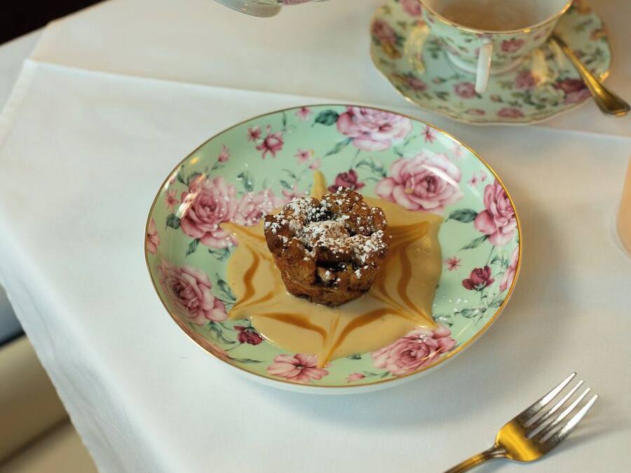 Eine blumengeschmückte Teekanne gießt Tee in eine passende Tasse und Untertasse, ein Dessertteller und eine Gabel auf einem Tisch.