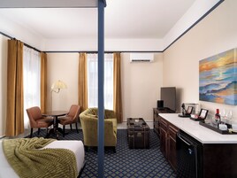 A hotel room with a bed, two chairs, a desk, a television, and a painting on the wall.