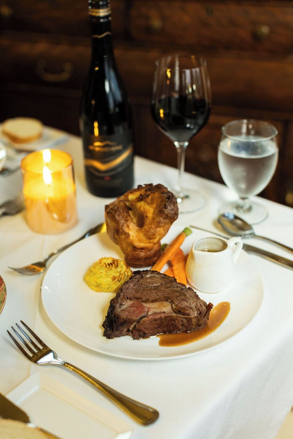 Une assiette contenant un steak, des Yorkshire puddings et des légumes, accompagnée de verres à vin et d’une bougie.