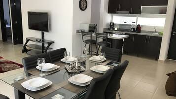 Un moderno comedor con una mesa de cristal, sillas negras y platos blancos. Se observa una cocina al fondo con armarios oscuros y una estufa.