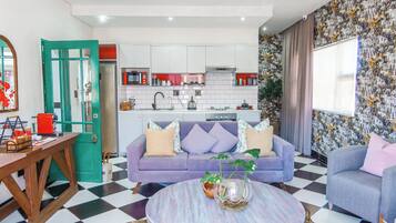 A modern living room with a purple sofa, a round coffee table, and a kitchen in the background.