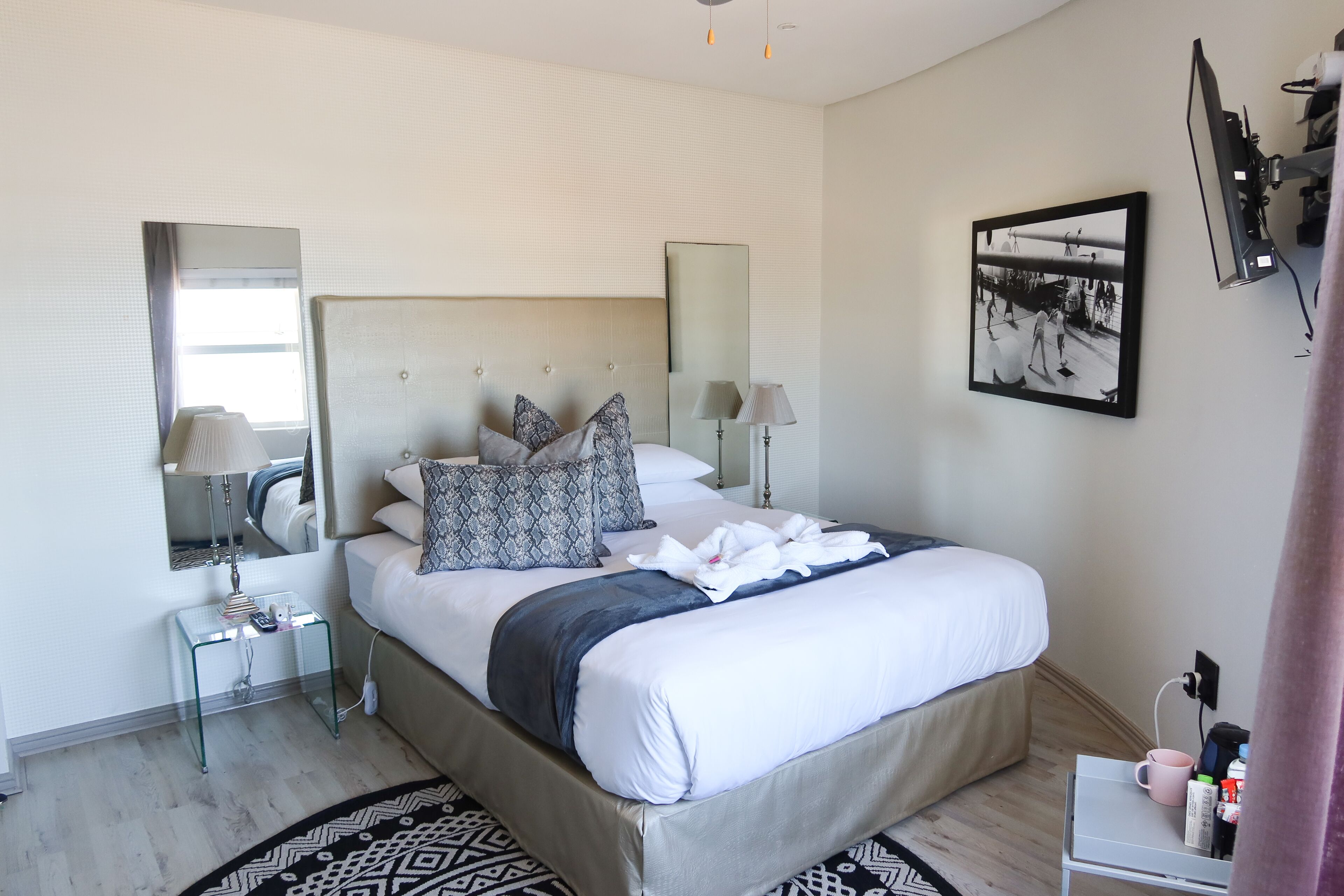 A modern hotel room with a large bed, bedside tables, a mirror, and a framed photograph on the wall.