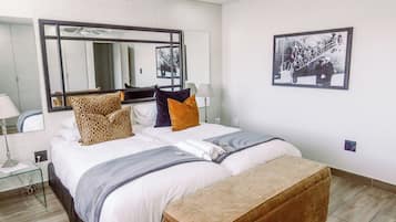 A hotel room with a large bed, a bench, a mirror, a lamp, and a framed picture on the wall.