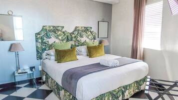 A hotel room with a bed, a headboard with a leaf pattern, a mirror, a lamp, and a window with curtains.