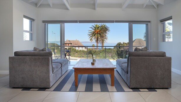 Una sala de estar moderna con un gran ventanal corredizo que da a un balcón con vistas al mar y una palmera. Hay dos sofás grises y una mesa de centro de madera en la habitación.