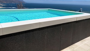Piscina rectangular con agua azul cristalina, bordeada por un borde de baldosas blancas, frente a un paisaje costero montañoso y un cielo despejado.