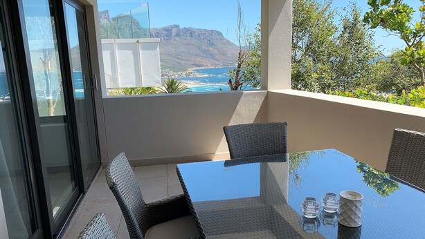 Un balcón con mesa de vidrio y sillas de mimbre, con vistas a un paisaje costero con montañas y una playa.