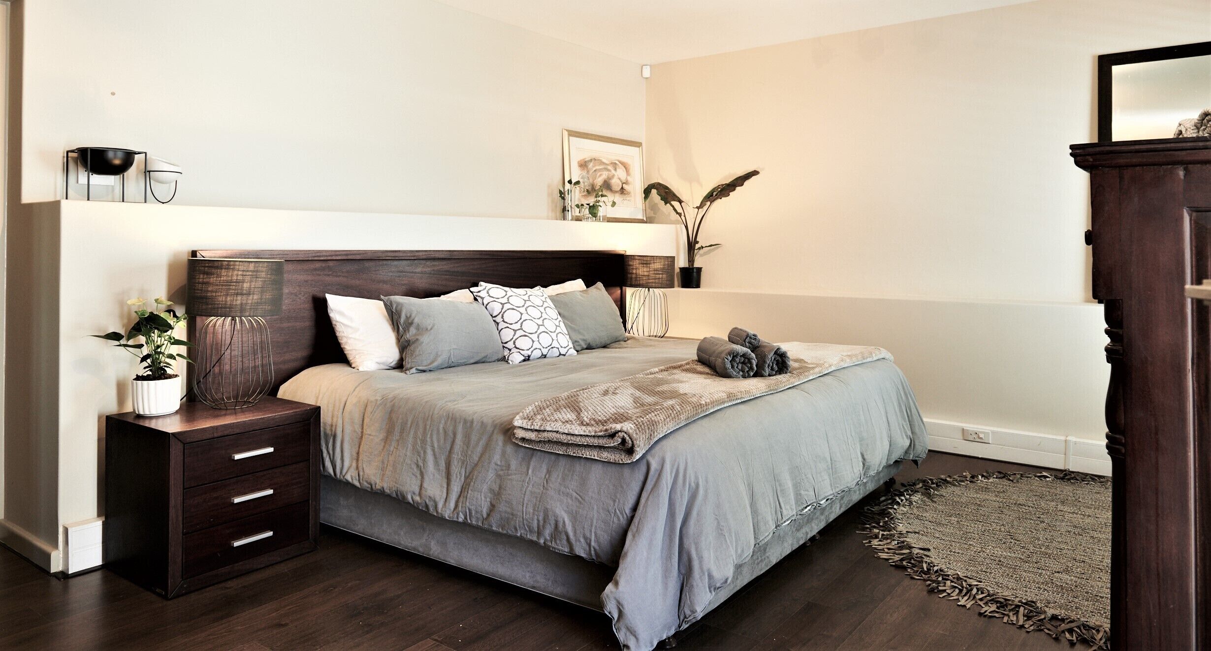 Une chambre à coucher avec un lit, des tables de chevet, une commode, une lampe, un miroir, une plante et un tableau au mur.