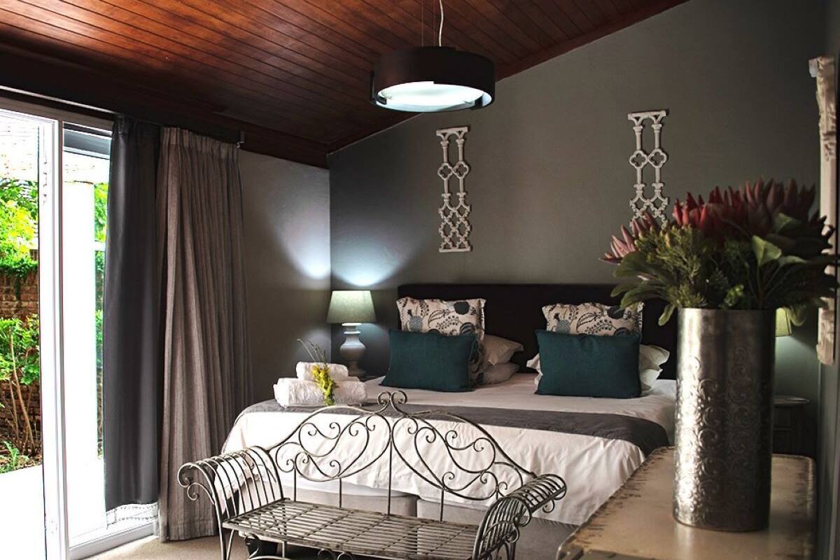 A bedroom with a bed, a metal bench, a vase with flowers, and a view of greenery outside.