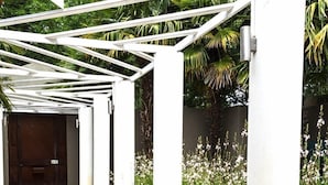 A covered walkway with white columns and a tiled floor, surrounded by greenery and flowers. - Kroon Manor (Moqhaka)