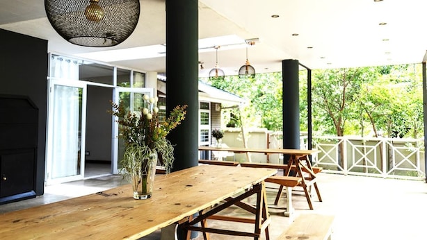 A wooden dining table with benches, a vase with flowers, and hanging pendant lights in a modern interior with large windows and a view of greenery outside. - Kroon Manor (Moqhaka)