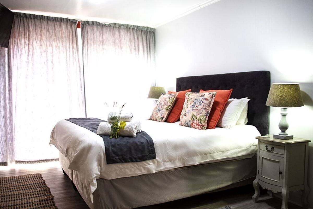A neatly made bed with white linens, a dark headboard, and multiple pillows. There is a bedside table with a lamp and a small drawer.