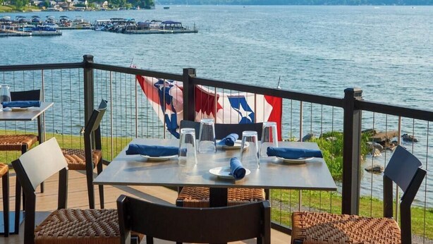 Een terras met tafels en stoelen, met uitzicht op een meer met boten en een steiger op de achtergrond.