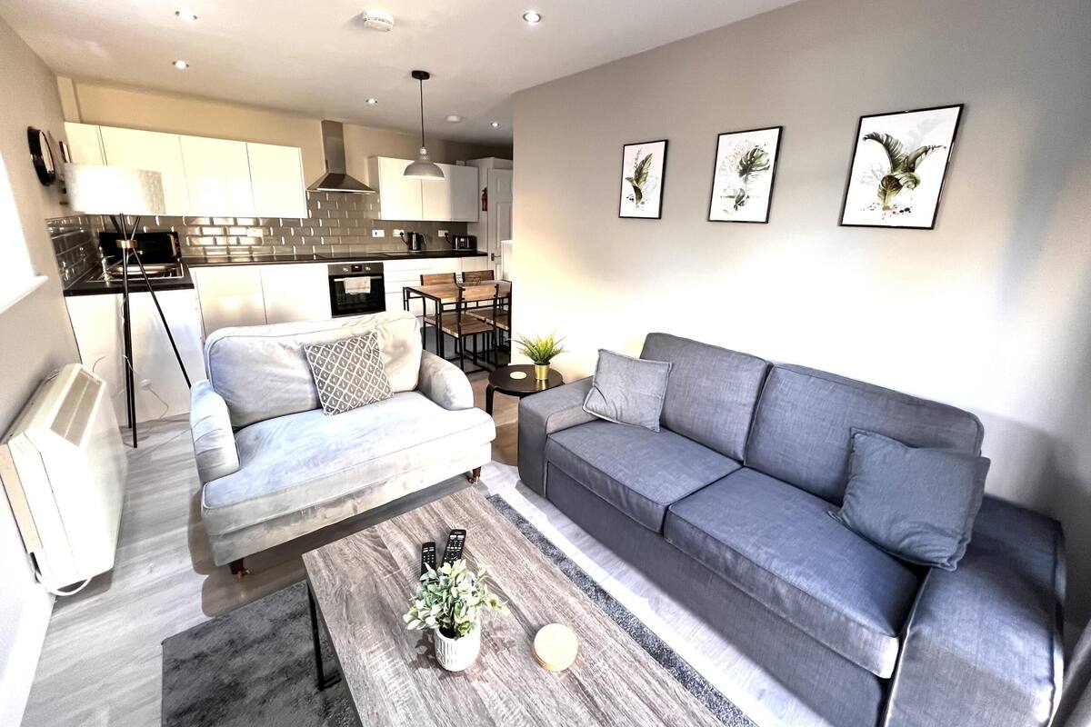 A modern living room with a grey sofa, a coffee table with a plant, and a kitchen in the background.