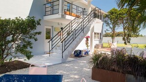 Un bâtiment moderne à deux étages, doté d’un escalier menant à un balcon, entouré de verdure et agrémenté d’un espace de détente extérieur.