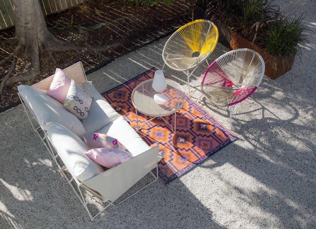 Un espace extérieur aménagé avec un canapé blanc, deux chaises en métal, un tapis à motifs et un bac à fleurs contenant des plantes vertes.
