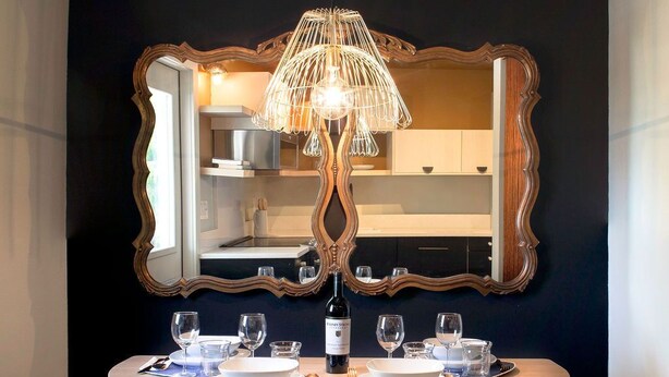 Un coin repas avec une table ronde, deux tabourets bleus rembourrés, une bouteille de vin et un miroir doté d’un cadre orné.