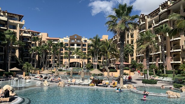 Ein Poolbereich eines Resorts mit mehreren Gebäudebereichen, Palmen und Liegestühlen.