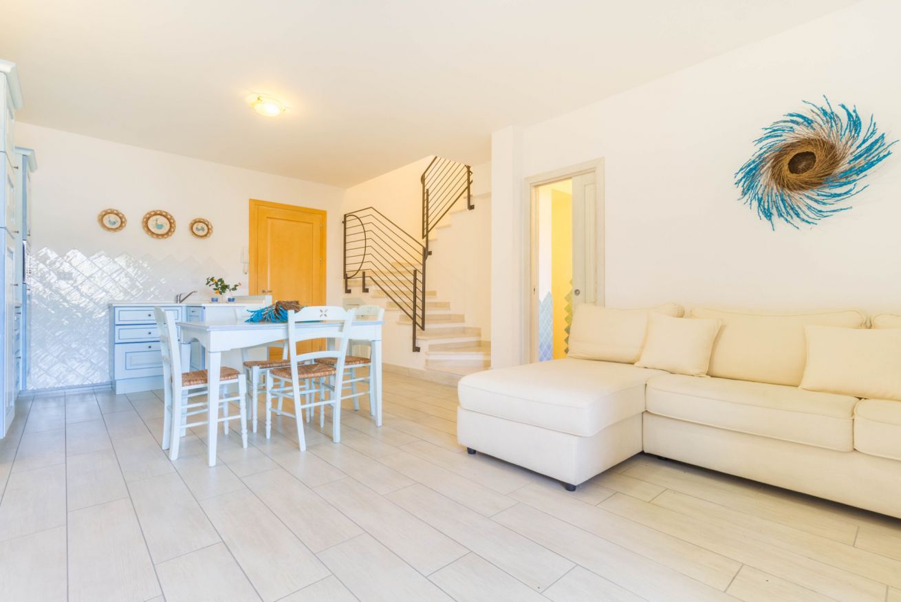 A modern living room with a white sofa, a dining table with chairs, a staircase, and a decorative wall piece.
