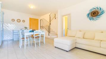 A modern living room with a white sofa, a dining table with chairs, a staircase, and a decorative wall piece.