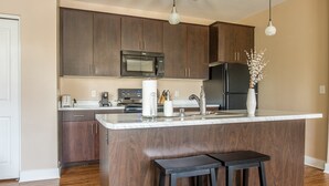 A modern kitchen with dark wood cabinets, a stainless steel refrigerator, a microwave, and a white marble island with two stools. - Sandia Peak Apartments by Frontdesk (Albuquerque)