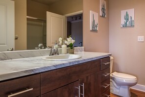 A bathroom with a marble sink, dark wooden cabinets, a white toilet, and cactus-themed wall art. - Sandia Peak Apartments by Frontdesk (Albuquerque)