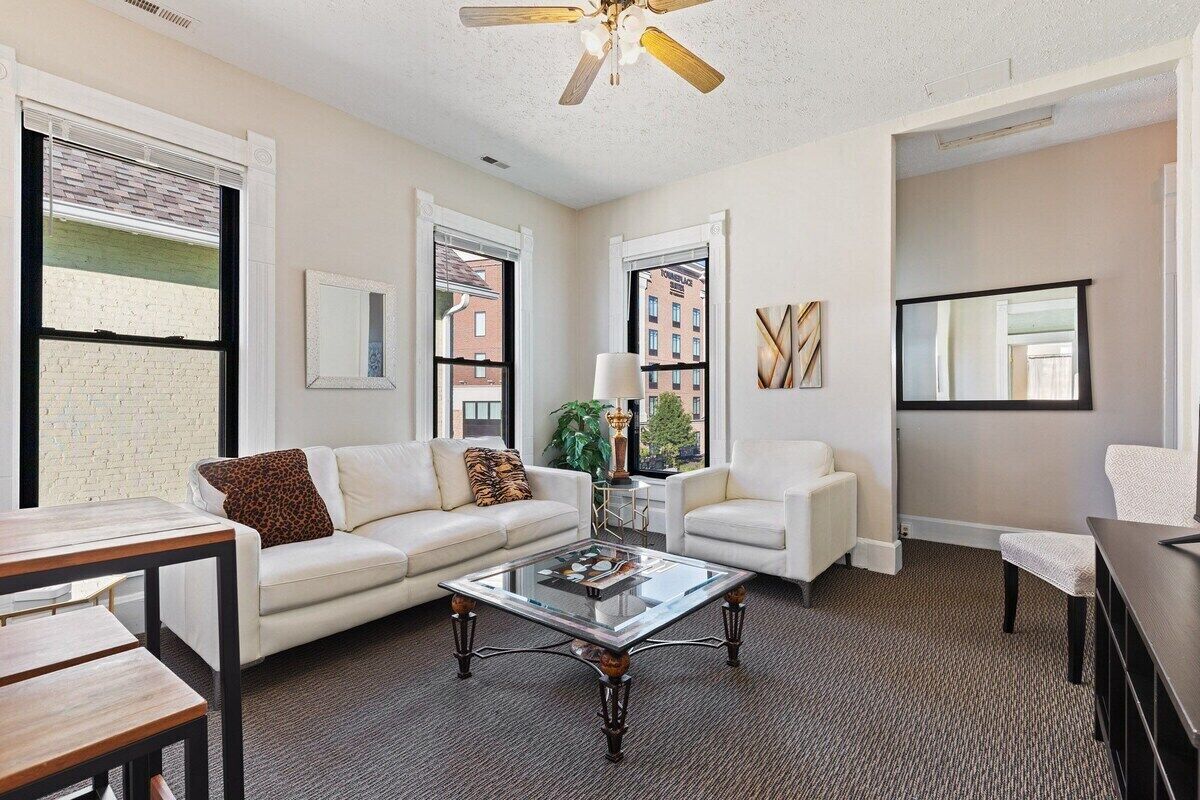 A modern living room with a glass coffee table, white sofas, and a ceiling fan.