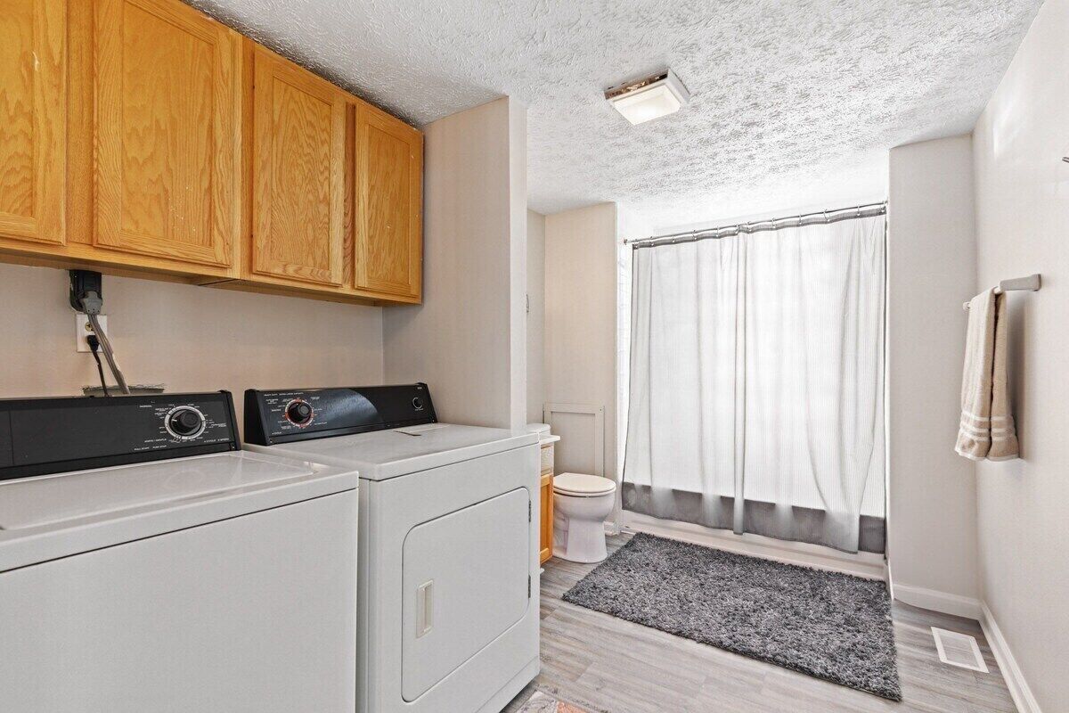 A laundry area with two washing machines, wooden cabinets, a window with curtains, and a tiled floor.