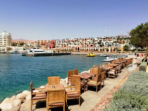 A waterfront restaurant with outdoor seating, a marina with boats, and colorful buildings in the background. - Three Bedroom Flats 8 (Aqaba)