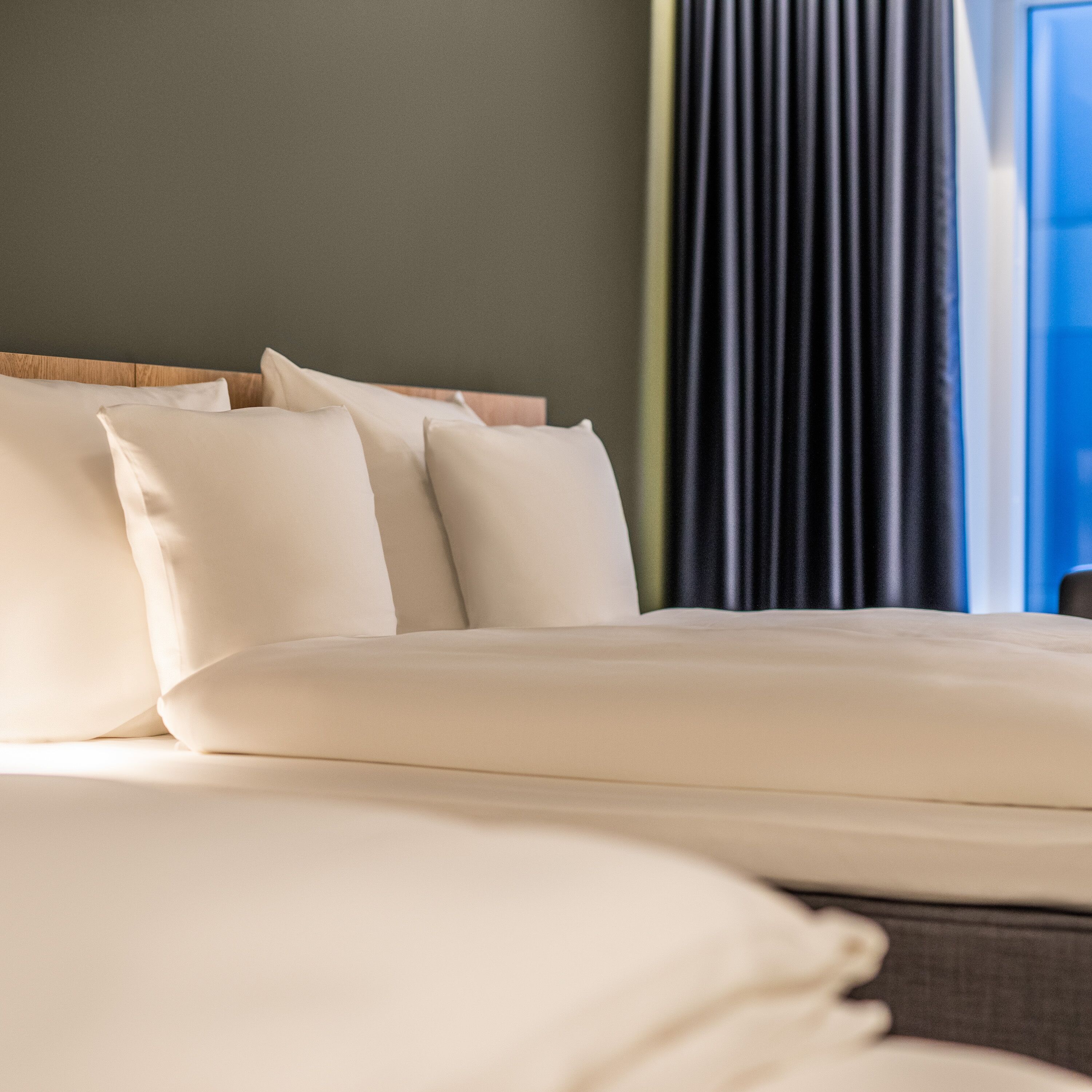 Una cama bien hecha con ropa de cama y almohadas blancas, un cabecero de madera y una ventana con cortinas oscuras.
