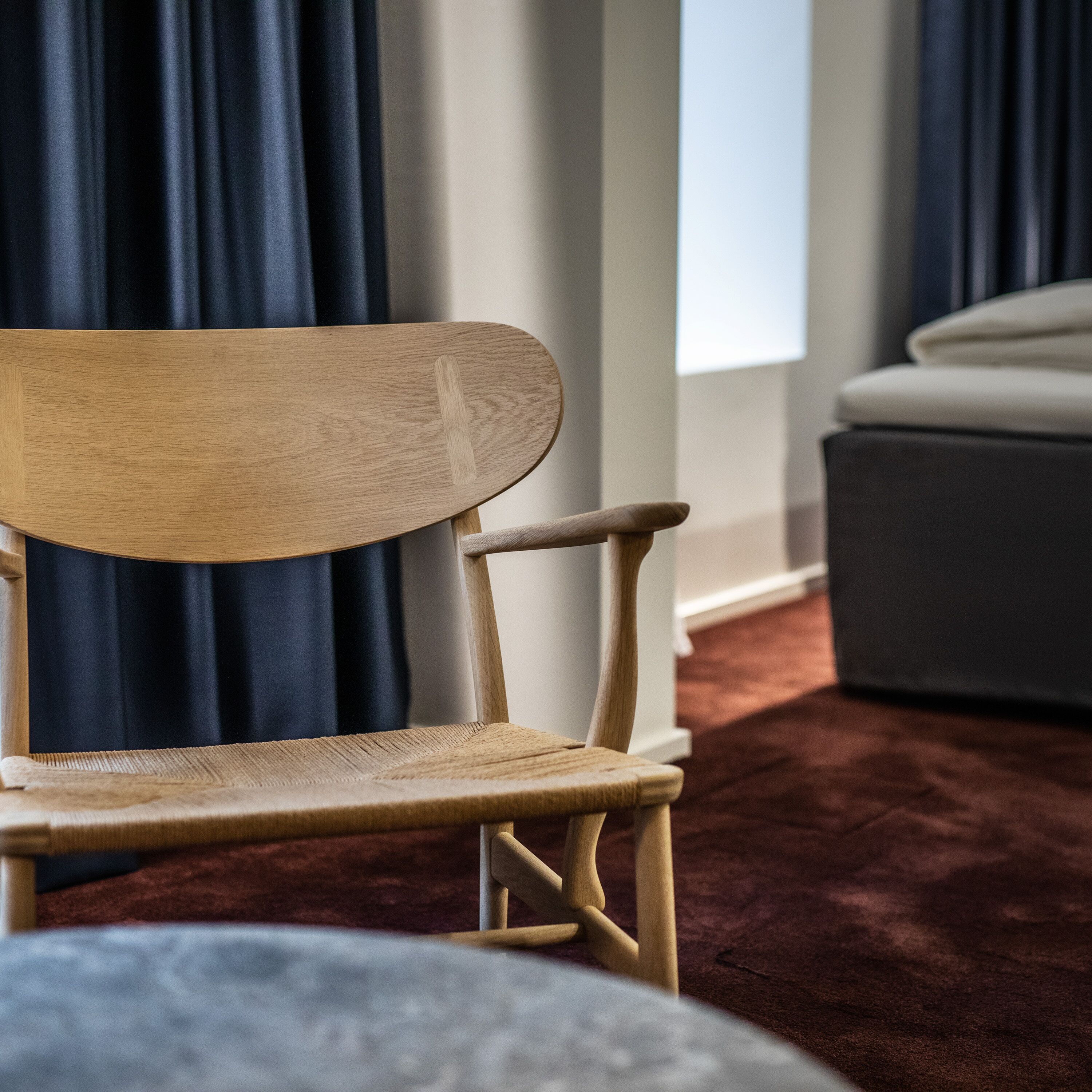 A wooden chair with a curved backrest and armrests, positioned in a room with a dark curtain and a light-colored sofa in the background.