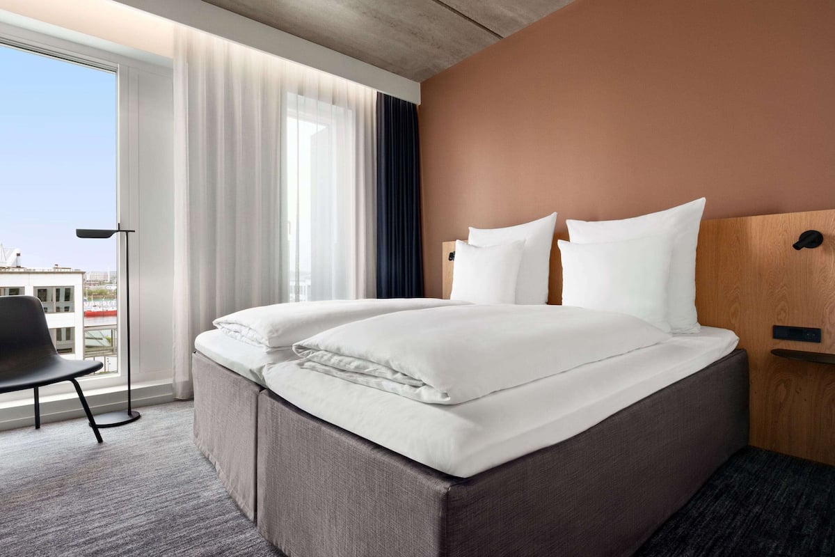 Quarto de hotel com uma cama grande, uma cadeira e vista para a cidade pela janela.